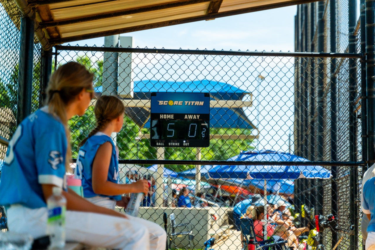 Two softball girls looking at a blue Score Titan scoreboard in the dugout